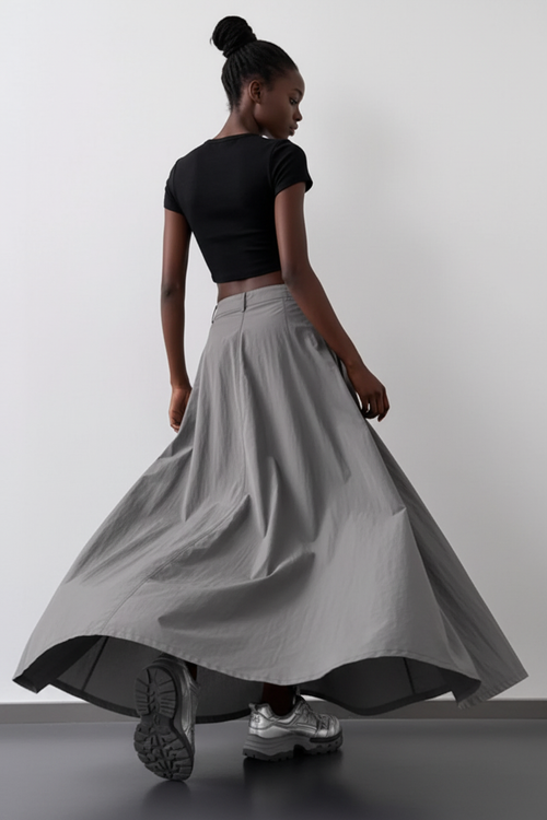  Jupe Maxi Cargo Grise – Volume et Attitude de Dos
 Vue de dos d'une mannequin portant une jupe maxi cargo grise en tissu nylon froissé en mouvement dramatique sur fond blanc — silhouette évasée A-line volumineuse, tissu crinkle texturé aux reflets métalliques subtils, taille basse avec passants de ceinture, portée avec un crop top noir et sneakers argentées. Look streetwear audacieux et féminin.