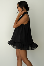 Robe mini halter Black-Coat noire avec double étage de volants - profil latéral complet d'une mannequin en mouvement gracieux sur fond blanc studio, soulignant la silhouette élancée créée par la coupe halter, le contraste entre le haut ajusté et la jupe volantée, et l'élégance intemporelle du noir profond
